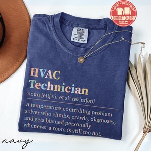 May include: Navy blue Comfort Colors t-shirt with the text "HVAC Technician" in colorful letters. The definition of HVAC Technician is printed below the title. A gold necklace with a pendant is draped on the shirt.