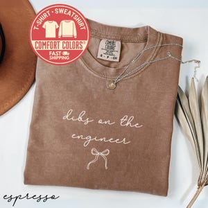 May include: A brown Comfort Colors t-shirt with the phrase "dibs on the engineer" in white script. A silver necklace with a pendant rests on the shirt. The shirt has a crew neck and short sleeves. A red and white circular logo is in the upper left corner.