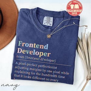 May include: Navy blue t-shirt with the text "Frontend Developer" in colorful letters. The definition of "Frontend Developer" is printed below the title. A gold necklace and a brown hat are also in the image.