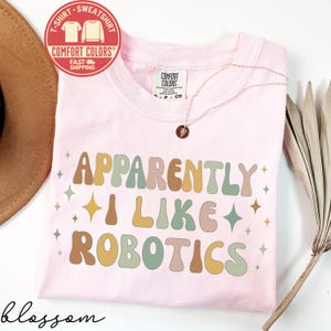 May include: A light pink t-shirt with the text "APPARENTLY I LIKE ROBOTICS" in retro-style lettering. The shirt has a crew neck and short sleeves. A gold necklace with a pendant is visible. A red and white Comfort Colors logo is in the upper left corner.