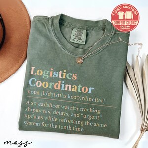 May include: Olive green t-shirt with the text "Logistics Coordinator" in colorful letters. The shirt includes a definition and a description of the job. A gold necklace and a brown hat are also visible.