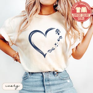 May include: An ivory short-sleeved t-shirt featuring a dark blue graphic of a heart outline. The heart design incorporates a ballet slipper, a musical note, two small four-pointed stars, and an infinity symbol along its right-hand curve.
