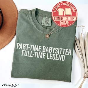 May include: An olive green t-shirt with the text "PART-TIME BABYSITTER FULL-TIME LEGEND" in white. The shirt has a crew neck and short sleeves. A gold necklace and a Comfort Colors logo are also visible.