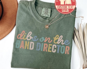 May include: An olive green t-shirt with the phrase "dibs on the BAND DIRECTOR" in colorful letters. A gold necklace with a pendant rests on the shirt. The shirt has a Comfort Colors label.