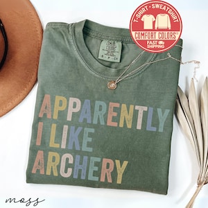 May include: An olive green t-shirt with the text "APPARENTLY I LIKE ARCHERY" in pastel colors. The shirt has a crew neck and short sleeves. A gold necklace and a brown hat are also visible.