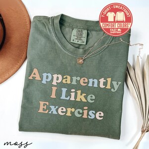 May include: An olive green t-shirt with the phrase "Apparently I Like Exercise" in pastel colors. The shirt has a crew neck and a Comfort Colors tag. A gold necklace and a dried plant are also visible.