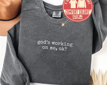 May include: A dark gray Comfort Colors sweatshirt with the text "god's working on me, ok?" in white. The sweatshirt has a crew neck and long sleeves. A brown hat and a gold necklace are also visible.