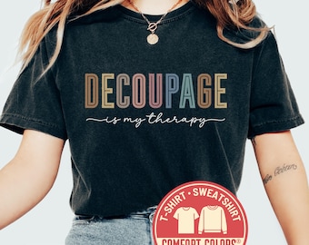 May include: Black t-shirt with the text "DECOUPAGE is my therapy" in colorful letters. The shirt has a round neck and short sleeves. A red circular logo with the words "T-SHIRT SWEATSHIRT" and "COMFORT COLORS" is visible. The word "black" is written on the bottom left.