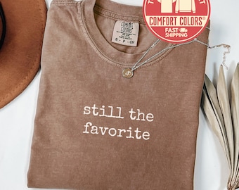 May include: Espresso-colored Comfort Colors t-shirt with the phrase "still the favorite" in white. A silver necklace with a pendant rests on the shirt. The shirt has a crew neck and short sleeves. A brown hat and dried plant are in the background.