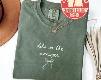 May include: An olive green t-shirt with the phrase "dibs on the manager" in white script, accompanied by a bow graphic. The shirt features a Comfort Colors label and a round red sticker that says "T-shirt Sweatshirt Comfort Colors Fast Shipping". A gold necklace is draped on the shirt.