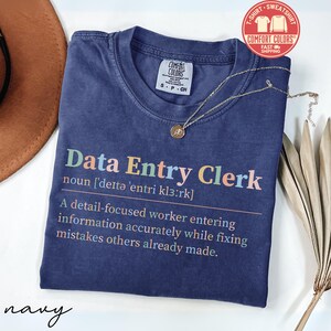 May include: Navy blue Comfort Colors t-shirt with the text "Data Entry Clerk" in colorful letters. The definition of the job is printed below. A gold necklace with a pendant is on the shirt.