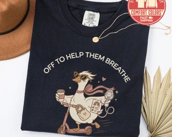 May include: A black t-shirt featuring a graphic of a goose wearing sunglasses and a scarf, riding a scooter while holding a coffee cup and a medical nebulizer. The text reads OFF TO HELP THEM BREATHE. A gold chain necklace rests on the collar.