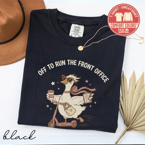 May include: A black crewneck t-shirt featuring a graphic of a goose wearing sunglasses and a scarf, riding a scooter while holding a coffee cup and a clipboard. The text reads OFF TO RUN THE FRONT OFFICE. A gold chain necklace rests on the shirt.