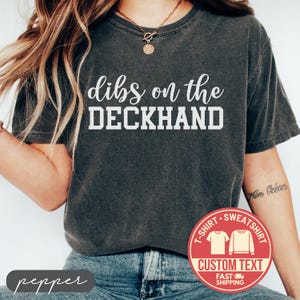 May include: Dark gray t-shirt with the text "dibs on the DECKHAND" in white. The shirt has a crew neck and short sleeves. A red circular graphic with the words "CUSTOM TEXT" is visible.