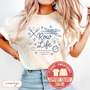 May include: Ivory-colored t-shirt with the text "Row Life" in navy blue, featuring oars, boats, stars, and a stopwatch graphic. The shirt has a crew neck and short sleeves. A red Comfort Colors logo is visible.