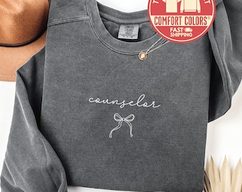 Puede incluir: Una sudadera Comfort Colors gris oscuro con la palabra "counselor" en cursiva y un lazo gráfico. La sudadera tiene cuello redondo y mangas largas. Se ve un collar con un colgante.