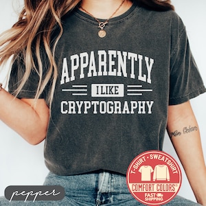May include: Dark gray t-shirt with the text "APPARENTLY I LIKE CRYPTOGRAPHY" in white block letters. The shirt has a crew neck and short sleeves. A red and white logo is visible at the bottom right.