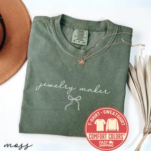 May include: An olive green t-shirt with the words "jewelry maker" in white script and a bow graphic. A gold necklace with a pendant rests on the shirt. A brown hat and dried plant are in the background.