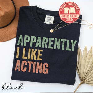 May include: Black crew neck t-shirt with the text "APPARENTLY I LIKE ACTING" in green, yellow, and orange. The shirt has a Comfort Colors tag and a red sticker that says "T-Shirt Sweatshirt" and "Fast Shipping". A gold necklace and a brown hat are also visible.
