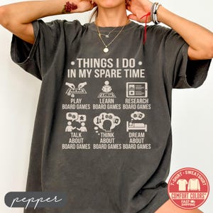 May include: Dark gray Comfort Colors t-shirt with the text "THINGS I DO IN MY SPARE TIME" and illustrations of board game activities. Includes "PLAY", "LEARN", "RESEARCH", "TALK ABOUT", "THINK ABOUT", and "DREAM ABOUT" board games.