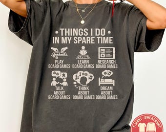 May include: Dark gray Comfort Colors t-shirt with the text "THINGS I DO IN MY SPARE TIME" and illustrations of board game activities. Includes "PLAY", "LEARN", "RESEARCH", "TALK ABOUT", "THINK ABOUT", and "DREAM ABOUT" board games.
