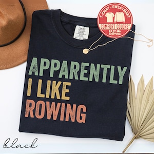 May include: A black t-shirt with the text "APPARENTLY I LIKE ROWING" in green, yellow, and orange. The shirt has a crew neck and a Comfort Colors tag. A gold necklace and a brown hat are also visible.