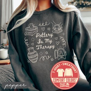 May include: A dark gray sweatshirt with the phrase "Pottery Is My Therapy" in white script, along with white line drawings of pottery tools and a kiln. The sweatshirt has a crew neck and long sleeves. The brand name "Comfort Colors" is visible.