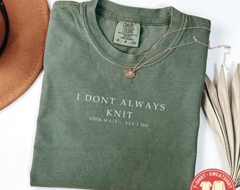 May include: A moss green short-sleeved t-shirt with a crew neckline. The shirt features the white printed text I DONT ALWAYS KNIT OHH WAIT... YES I DO. A thin gold-toned necklace with a small circular pendant rests on the fabric near the collar.