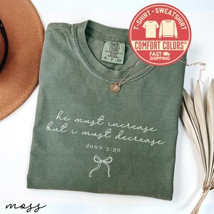 May include: An olive green t-shirt with the text "he must increase but I must decrease" and "JOHN 3:30" printed in white. A gold necklace with a pendant rests on the shirt. The shirt has a Comfort Colors label.
