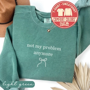 May include: A light green Comfort Colors sweatshirt with the phrase "not my problem anymore" in white script. A small bow graphic is below the text. The shirt has a crew neck and long sleeves. A round sticker with the words "T-Shirt Sweatshirt" is in the upper right corner.