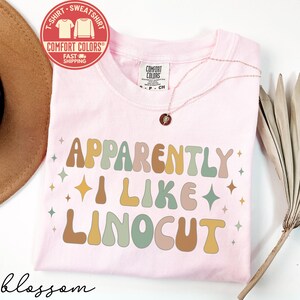 May include: A light pink t-shirt with the text "APPARENTLY I LIKE LINOCUT" in retro-style lettering. The shirt has a crew neck and short sleeves. A gold necklace with a pendant is on the shirt. A red and white sticker is on the top left.