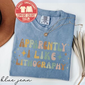 May include: A blue jean Comfort Colors t-shirt with the text "Apparently I Like Lithography" in colorful retro font. The shirt has a crew neck and short sleeves. A gold necklace with a pendant is draped on the shirt.