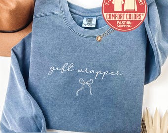 May include: Blue jean colored sweatshirt with the words "gift wrapper" in white script and a bow graphic. The sweatshirt has a crew neck and long sleeves. A brown hat is partially visible in the background.