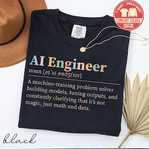 May include: Black t-shirt with the text "AI Engineer" in colorful letters. The shirt includes a definition of the term, and the brand name "Comfort Colors". A gold necklace and a brown hat are also visible.