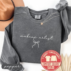 May include: A dark gray Comfort Colors sweatshirt with the words "makeup artist" in white script and a bow graphic. A gold necklace and a brown hat are visible. The sweatshirt has a crew neck and a red and white Comfort Colors logo.