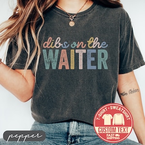 May include: Dark gray t-shirt with the phrase "dibs on the WAITER" in colorful, distressed lettering. A red circular graphic with "T-SHIRT SWEATSHIRT CUSTOM TEXT FAST SHIPPING" is visible. The shirt is a casual wear item.