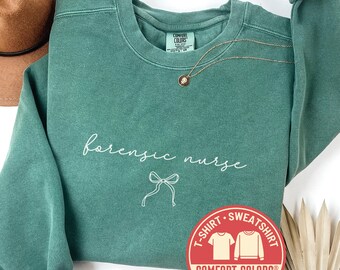 May include: Light green sweatshirt with the words "forensic nurse" in white script and a bow graphic. A gold necklace with a pendant is on the sweatshirt. The Comfort Colors brand logo is visible. A brown hat is in the background.