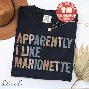 May include: Black t-shirt with the text "APPARENTLY I LIKE MARIONETTE" in colorful block letters. A gold necklace and a brown hat are visible. The shirt has a Comfort Colors tag and a red sticker that says "T-SHIRT SWEATSHIRT".