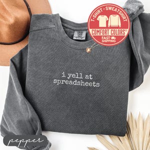 May include: Pepper-colored Comfort Colors sweatshirt with the embroidered phrase "i yell at spreadsheets." The crew neck sweatshirt has a relaxed fit and a vintage wash. A red and white circular graphic with the words "T-shirt Sweatshirt" is visible.