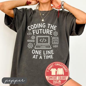 May include: Dark grey t-shirt with the text "CODING THE FUTURE" and a laptop graphic. The shirt also features binary code and the phrase "ONE LINE AT A TIME". A red and white Comfort Colors logo is visible.