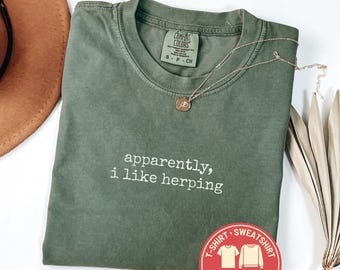 May include: A moss green Comfort Colors t-shirt with the text "apparently, i like herping" in white. A gold-colored necklace with a pendant rests on the shirt. A brown hat and a Comfort Colors logo are also visible.