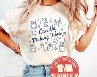 Puede incluir: Camiseta color crema con el texto "Candle Making Vibes" en azul, rodeado de ilustraciones de velas. La camiseta presenta un logotipo de Comfort Colors y es una camiseta o sudadera.