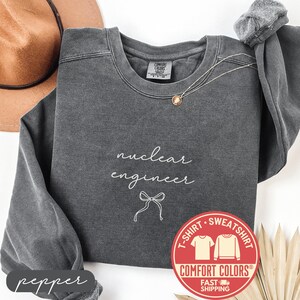 May include: A pepper-colored Comfort Colors sweatshirt with the words "nuclear engineer" in white script, along with a bow graphic. A gold necklace and a brown hat are visible. A red and white Comfort Colors logo is on the right.