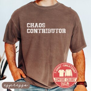 May include: Espresso-colored t-shirt with the text "CHAOS CONTRIBUTOR" in a distressed white font. The shirt features a Comfort Colors label and a fast shipping badge. The shirt is a t-shirt or sweatshirt.