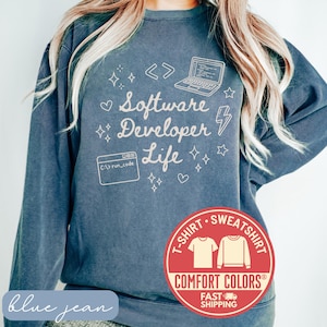 May include: Blue jean colored sweatshirt with the text "Software Developer Life" and graphics of a laptop, code symbols, and stars. A red circle with "T-Shirt Sweatshirt" and "Comfort Colors" is at the bottom.