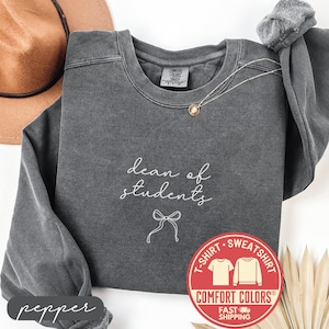 May include: A dark gray sweatshirt with the words "dean of students" in white script and a bow graphic. The sweatshirt has a crew neck and long sleeves. A Comfort Colors label is visible, along with a red and white circular graphic.