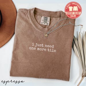May include: Espresso-colored t-shirt with the text "i just need one more tile" in white. The shirt has a crew neck and short sleeves. A silver necklace with a pendant is draped across the shirt. A Comfort Colors tag is visible.