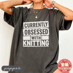 May include: A charcoal gray short-sleeve t-shirt featuring the white text CURRENTLY OBSESSED WITH KNITTING in a block-style font. The shirt has a crew neckline and a relaxed fit, with the text arranged in four distinct horizontal lines.