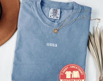 May include: A blue jean Comfort Colors t-shirt with the word "oma" embroidered in white. A gold necklace with a pendant rests on the shirt. A red sticker with the words "Custom Text" and "Fast Shipping" is on the bottom right.