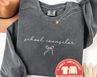 May include: A dark gray Comfort Colors sweatshirt with the words "school counselor" in white script and a bow graphic. A gold necklace and a brown hat are visible. The sweatshirt has a round red sticker that says "T-Shirt Sweatshirt Comfort Colors Fast Shipping".